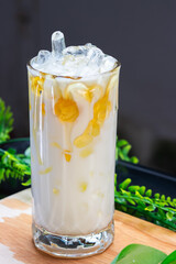 Chilled coconut milk drink with sweet corn and syrup drizzle, served with ice in a clear glass on a wooden surface and green background.