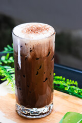 Iced chocolate drink topped with a layer of creamy foam and cocoa powder, served in a clear glass over ice on a wooden surface with green leaves.