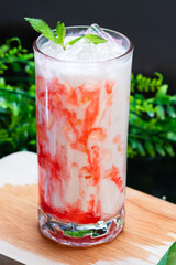 A vibrant iced strawberry milk drink swirled with red syrup, topped with fresh mint and ice, served in a clear glass on a wooden surface with greenery.