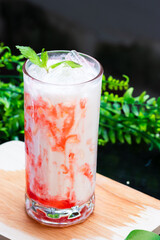 A vibrant iced strawberry milk drink swirled with red syrup, topped with fresh mint and ice, served in a clear glass on a wooden surface with greenery.