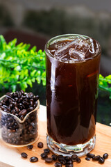 A refreshing iced black coffee in a tall glass with ice cubes, served alongside a clear container filled with roasted coffee beans on a wooden board.