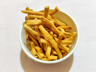 The white bowl contains the Indonesian cash food ladrang snack, which has a savory golden colored taste in the shape of a stick or pipe, served on a light colored cloth.