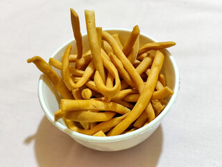 The white bowl contains the Indonesian cash food ladrang snack, which has a savory golden colored taste in the shape of a stick or pipe, served on a light colored cloth.
