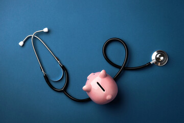 A stethoscope wrapped around a pink piggy bank on a blue background. Healthcare costs, savings and...