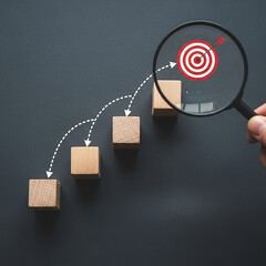 Photo of magnifying glass focusing on a target on wooden blocks representing steps towards business goals and achievement