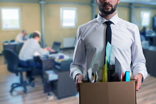 Office worker carrying a box of personal items, symbolizing job loss, resignation, or transition, with a blurred office background and coworkers at desks.