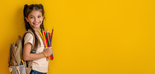 Smiling schoolgirl with a backpack holding colorful pencils against a yellow background, back to school