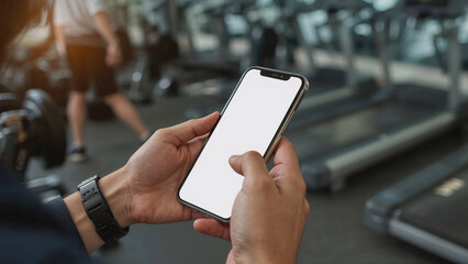 Businessman wearing a smartwatch and holding a smartphone with blank white screen in the gym, out of focus person exercising in the background. white screen copy space