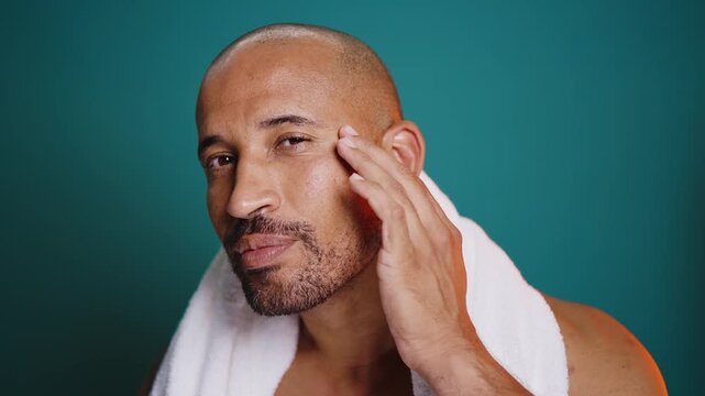 Attractive bald man with a white towel around his neck smiles showcasing a skincare routine against a brown backdrop