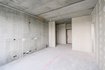 photo shows an unfinished apartment interior with bare concrete walls, ceiling, and floor. Plastered walls with visible joints, a door, and pipes