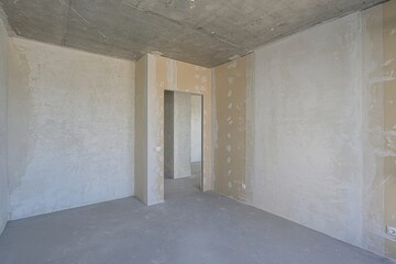 empty room under construction, with concrete ceiling and walls in rough plaster. There are doorways leading to other unfinished spaces