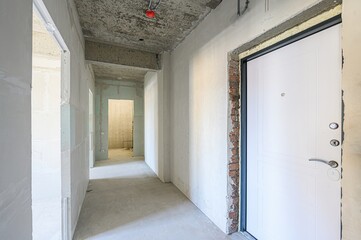 unfinished hallway with bare concrete floors and walls. A white door stands on the right, and open...