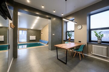 Modern interior with indoor pool view. Dining table with blue chairs next to large windows. Stylish and serene