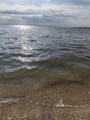 Sunny seascape with gentle waves lapping a sandy shore, distant reeds on the horizon, and shimmering reflections dancing on the water under a bright sky.
