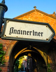 Obraz premium Old street sign pointing the way through a historic brick archway, with warm sunlight illuminating the ancient architecture and a glimpse of a charming urban alley beyond.