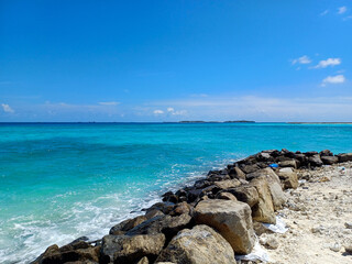 A rugged, rocky shoreline next to a vibrant turquoise ocean, with a distant island on the horizon and a bright blue sky. Related to tropical destinations, seaside vacation, natural coastal landscapes