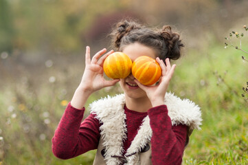 Playful Smiling girl sitting in grassy autumn field holding small pumpkins over her eyes, with a basket of apples beside her, celebrating Thanksgiving outdoors Autumn Harvest.