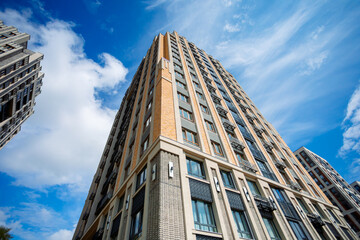 Fototapeta premium A modern residential complex with an elegant facade, made in light colors, with brick accents and large windows. Tall buildings, surrounded by blue sky and clouds, create a feeling of spaciousness and