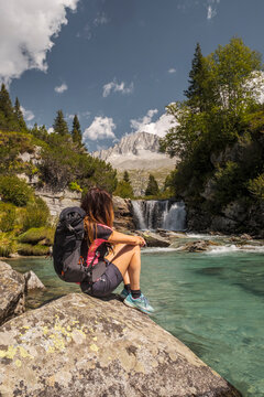 A woman with a backpack sits on a rock by a clear turquoise river, looking at a waterfall and mountain. Val di Fumo, Chiese river, Carr&egrave; Alto, Trentino. She is enjoying the scenic view during a hike.