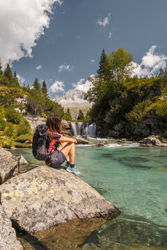A woman with a backpack sits on a rock by a clear turquoise river, looking at a waterfall and mountain. Val di Fumo, Chiese river, Carr&egrave; Alto, Trentino. She is enjoying the scenic view during a hike.