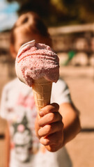 A child holding a pink ice cream cone toward the camera on a sunny summer day. The ice cream is in...