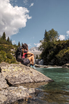 A woman with a backpack sits on a rock by a clear turquoise river, looking at a waterfall and mountain. Val di Fumo, Chiese river, Carr&egrave; Alto, Trentino. She is enjoying the scenic view during a hike.