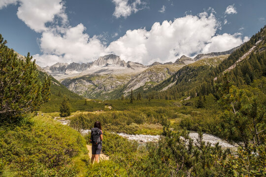 A woman with a backpack hikes towards mountains in a valley. Val di Fumo, Chiese river, Carr&egrave; Alto, Trentino. The scene shows a sunny day with some clouds in the sky.