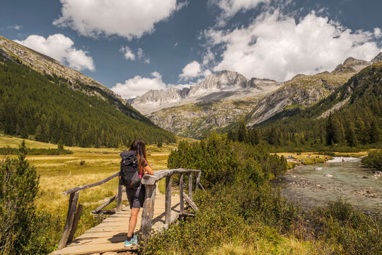 A hiker with a backpack stands on a wooden bridge, looking at a mountain landscape. Val di Fumo, Chiese river, Carr&egrave; Alto, Trentino. The scene captures the beauty of the Italian Alps on a sunny day.