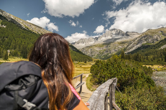 A woman with a backpack stands on a wooden bridge, looking at a mountain landscape with a river and trees. Val di Fumo, Chiese river, Carr&egrave; Alto, Trentino. 