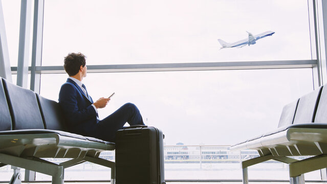 Young Businessman Sitting In Airport Departure Lounge Using Mobile Phone Watching Plane Take Off