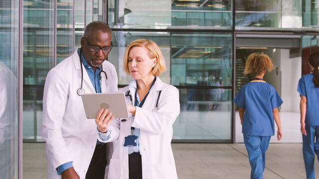 Male And Female Doctors Having Informal Meeting In Modern Hospital Looking At Digital Tablet
