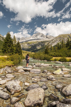 A hiker with a backpack stands on rocks in a stream, looking towards mountains under a cloudy sky. Val di Fumo, Chiese river, Carr&egrave; Alto, Trentino. The clear water flows around the rocks.