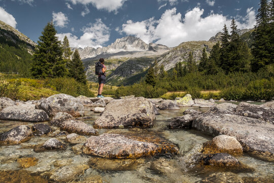 A hiker with a backpack stands on rocks in a stream, looking towards mountains under a cloudy sky. Val di Fumo, Chiese river, Carr&egrave; Alto, Trentino. The clear water flows around the rocks.