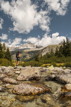 A hiker with a backpack stands on rocks in a stream, looking towards mountains under a cloudy sky. Val di Fumo, Chiese river, Carr&egrave; Alto, Trentino. The clear water flows around the rocks.