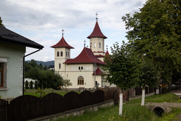 Fototapeta premium traditional orthodox monastery in a rural romanian village, featuring distinctive architecture, red-tiled roofs, and lush greenery under a cloudy sky