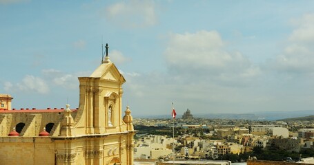 Santa Maria cathedral stands on island of Gozo near buildings and high flag of Malta. Landmark...