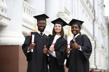 Portrait of a group of smiling happy multiracial international graduates students hugging and having fun in a university graduate gown and holding diploma outdoor. Education and graduation concept.