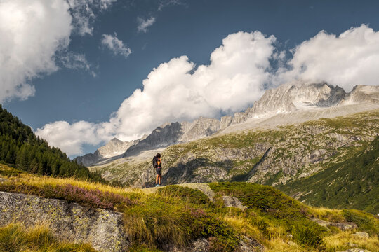 A hiker stands on a rocky outcrop overlooking a mountain valley with a river and bridge. Val di Fumo, Chiese river, Carr&egrave; Alto, Trentino.