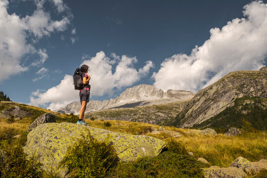 A hiker with a backpack stands on a mossy rock, gazing at a mountain range under a blue sky with scattered clouds. Val di Fumo, Chiese river, Carr&egrave; Alto, Trentino. 