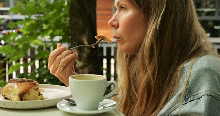 Mature woman with long loose blonde hair eats pastry dessert with fork and takes sip of coffee from white cup. Lady enjoys food and drink at outdoor setting