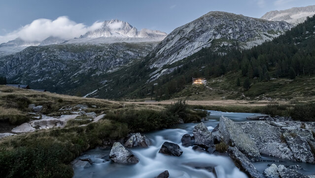 A scenic view of a mountain river flowing over rocks under a starry sky. Val di Fumo, Chiese river, Carr&egrave; Alto, Trentino. Lights from the Fumo Refuge are visible in the distance.
