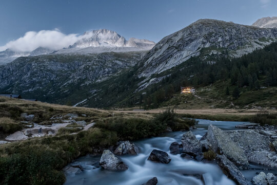 A scenic view of a mountain river flowing over rocks under a starry sky. Val di Fumo, Chiese river, Carr&egrave; Alto, Trentino. Lights from the Fumo Refuge are visible in the distance.