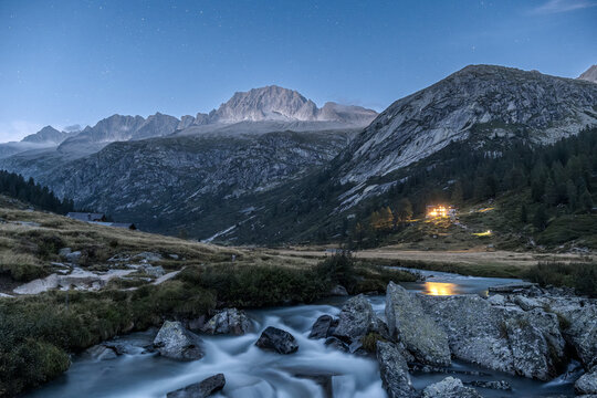 A scenic view of a mountain river flowing over rocks under a starry sky. Val di Fumo, Chiese river, Carr&egrave; Alto, Trentino. Lights from the Fumo Refuge are visible in the distance.