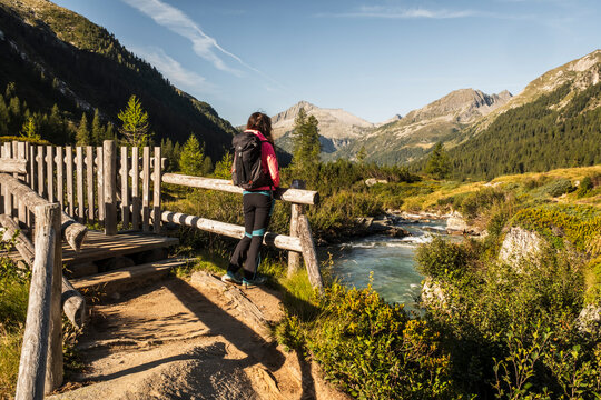 A hiker with a backpack pauses on a wooden bridge, gazing at a mountain river. Val di Fumo, Chiese river, Carr&egrave; Alto, Trentino. The scene is bathed in sunlight, highlighting the natural beauty.
