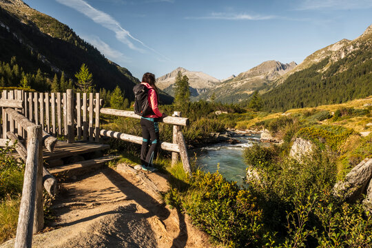 A woman with a backpack stands on a wooden bridge, looking out at a mountain river and valley. Val di Fumo, Chiese river, Carr&egrave; Alto, Trentino. She is enjoying the scenic view of the Italian Alps.