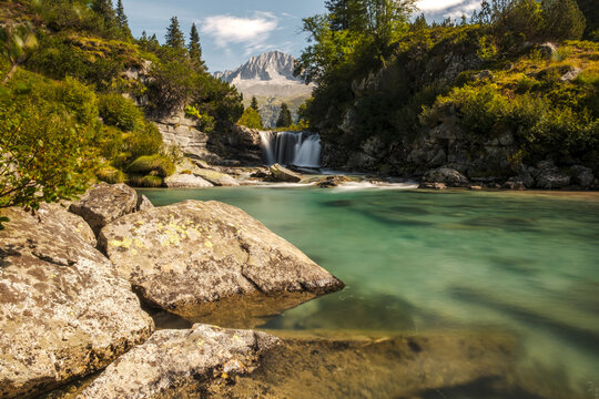 Val di Fumo, Chiese river, Carr&egrave; Alto, Trentino. A waterfall flows into a clear turquoise pool, surrounded by rocks and lush greenery, with a mountain in the background.