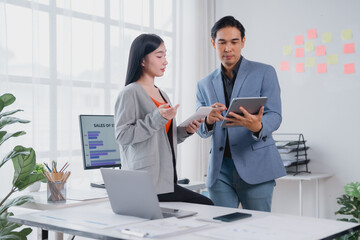 Two Asian business professionals engaging in a project discussion while utilizing a digital tablet and computer, collaborating effectively in a modern office environment