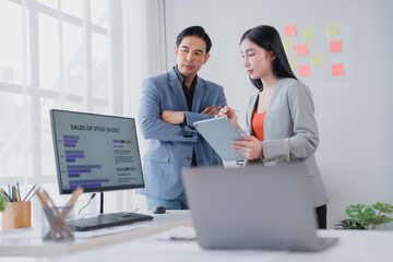 Two asian businesspeople discussing sales data on a tablet and desktop computer in a modern office,...