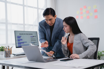 Two Asian business professionals engaging in a discussion about sales data in a bright office setting, utilizing a laptop, computer, and various documents for analysis and strategy planning