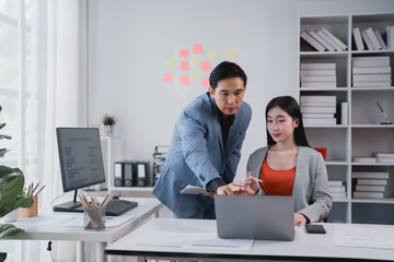 Two asian businesspeople are discussing work using a laptop in a bright office meeting,...